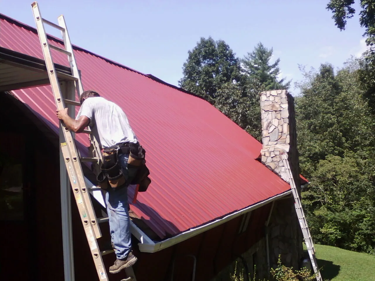 Roofer on a ladder beside a red metal roof and stone chimney — Soffit & Fascia Repair by Roofing Brothers in Wakefield